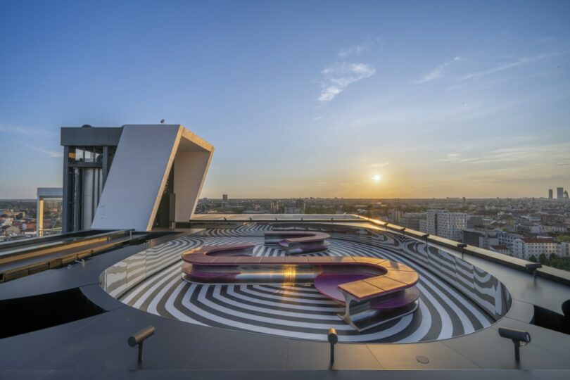 A rooftop with black-and-white striped circular patterns, sculptural purple benches, and a view of the city at sunset