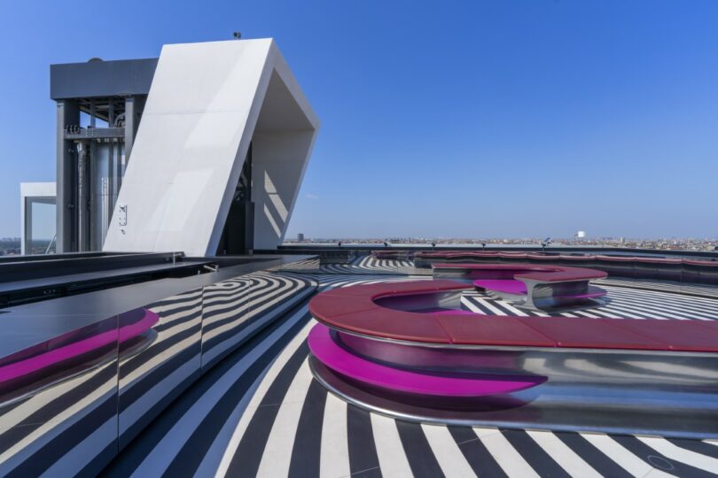 A modern rooftop terrace features black and white striped flooring, curved pink benches, and a large geometric white structure under a clear blue sky