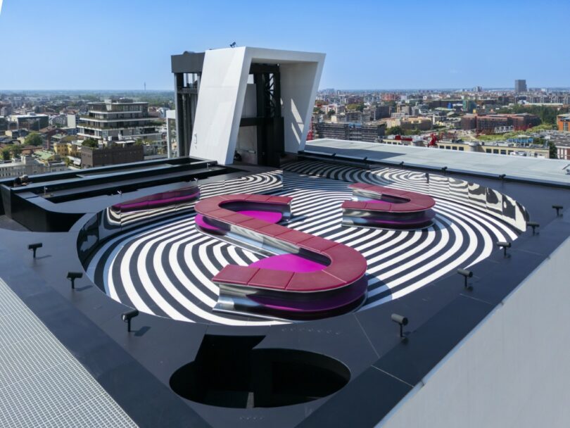 Rooftop with a large, pink S-shaped bench on top of a black and white spiraled pattern, overlooking a cityscape under a clear blue sky