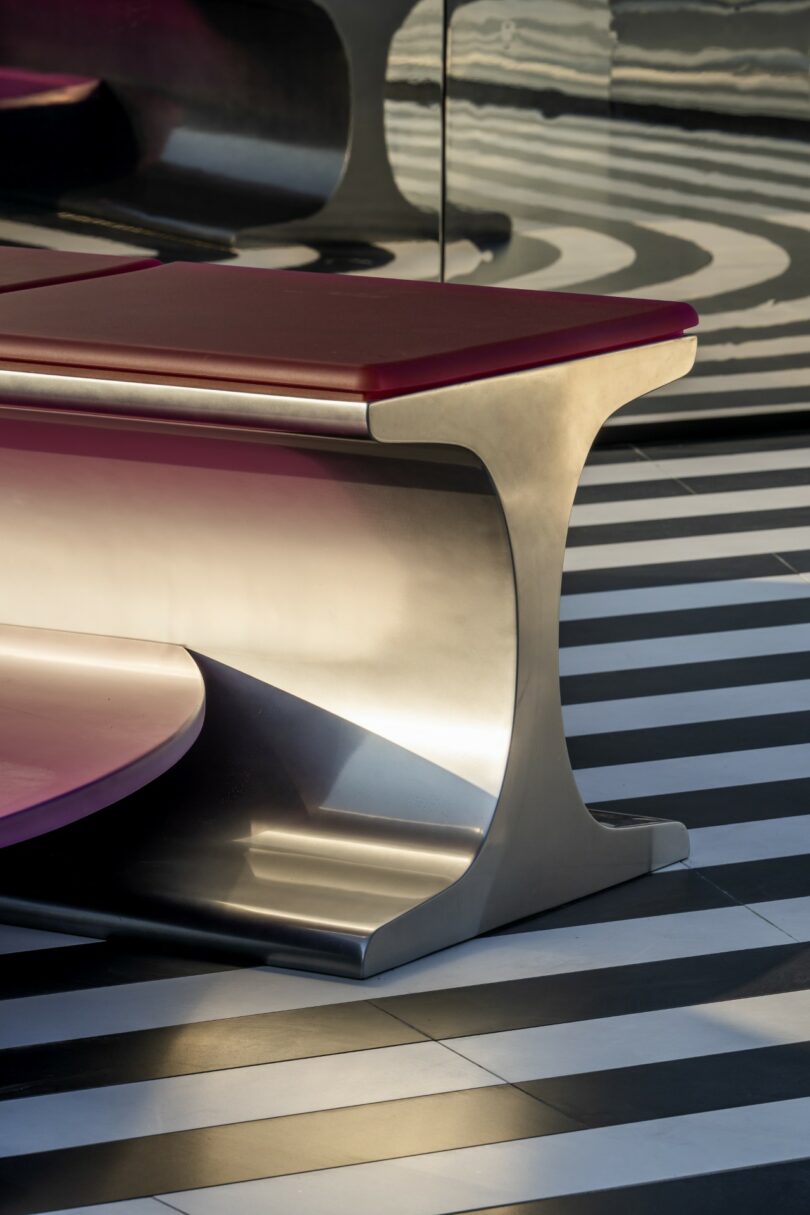 A modern bench with a metallic base and red cushion is placed on a black and white striped floor, with a reflective surface behind it
