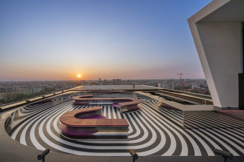 A rooftop terrace with striped black and white flooring, curved seating, and a view of the city at sunset