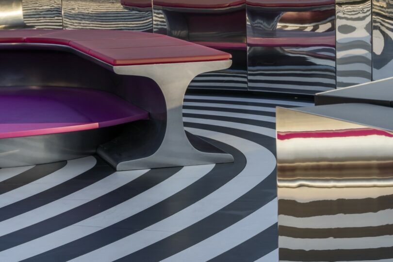 Curved bench with red and purple seating on a floor with black and white concentric circle patterns, next to reflective metallic surfaces