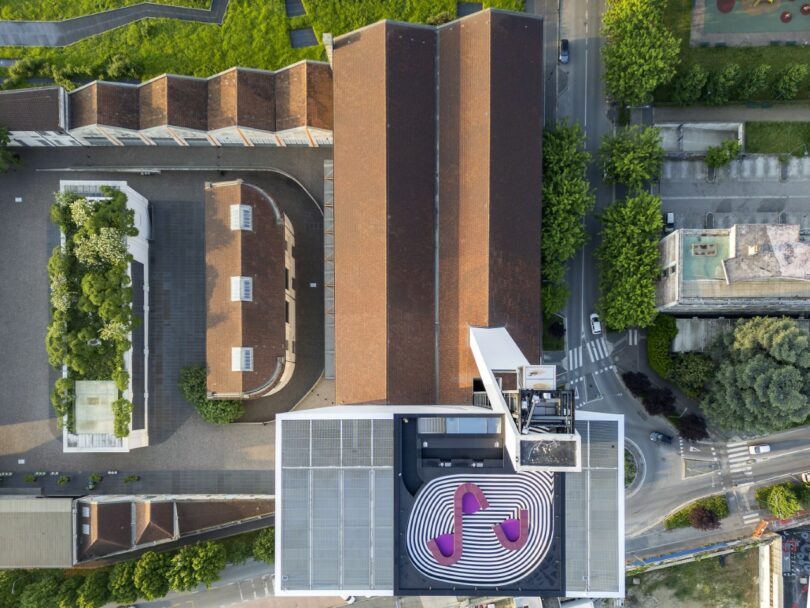 Aerial view of buildings with red roofs; one rooftop features a black and white swirl pattern with purple accents. Trees and a road surround the complex