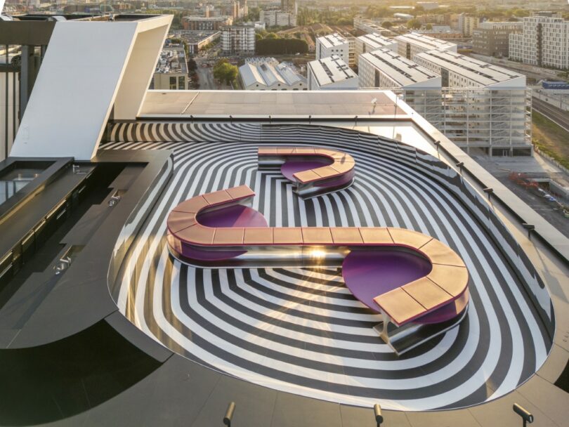 A rooftop area features curved wooden benches on a black and white striped pattern, surrounded by city buildings with an urban skyline in the background