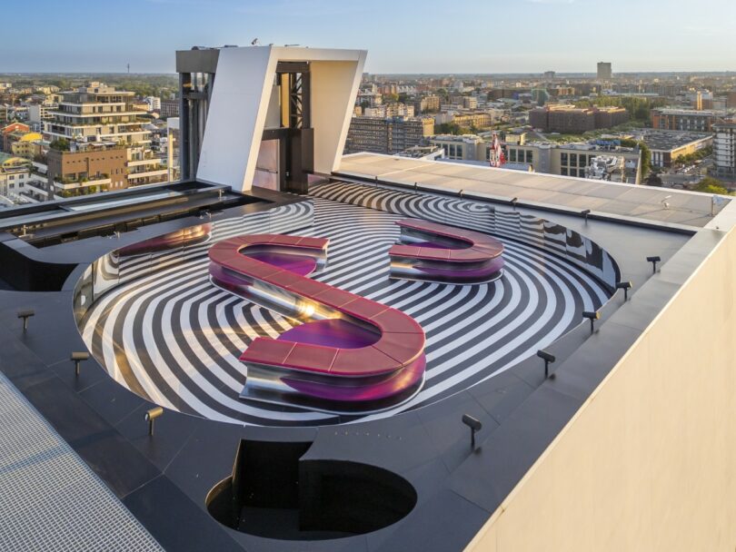 A rooftop area with black and white concentric circle patterns, featuring winding pink benches, overlooking a cityscape under a clear sky