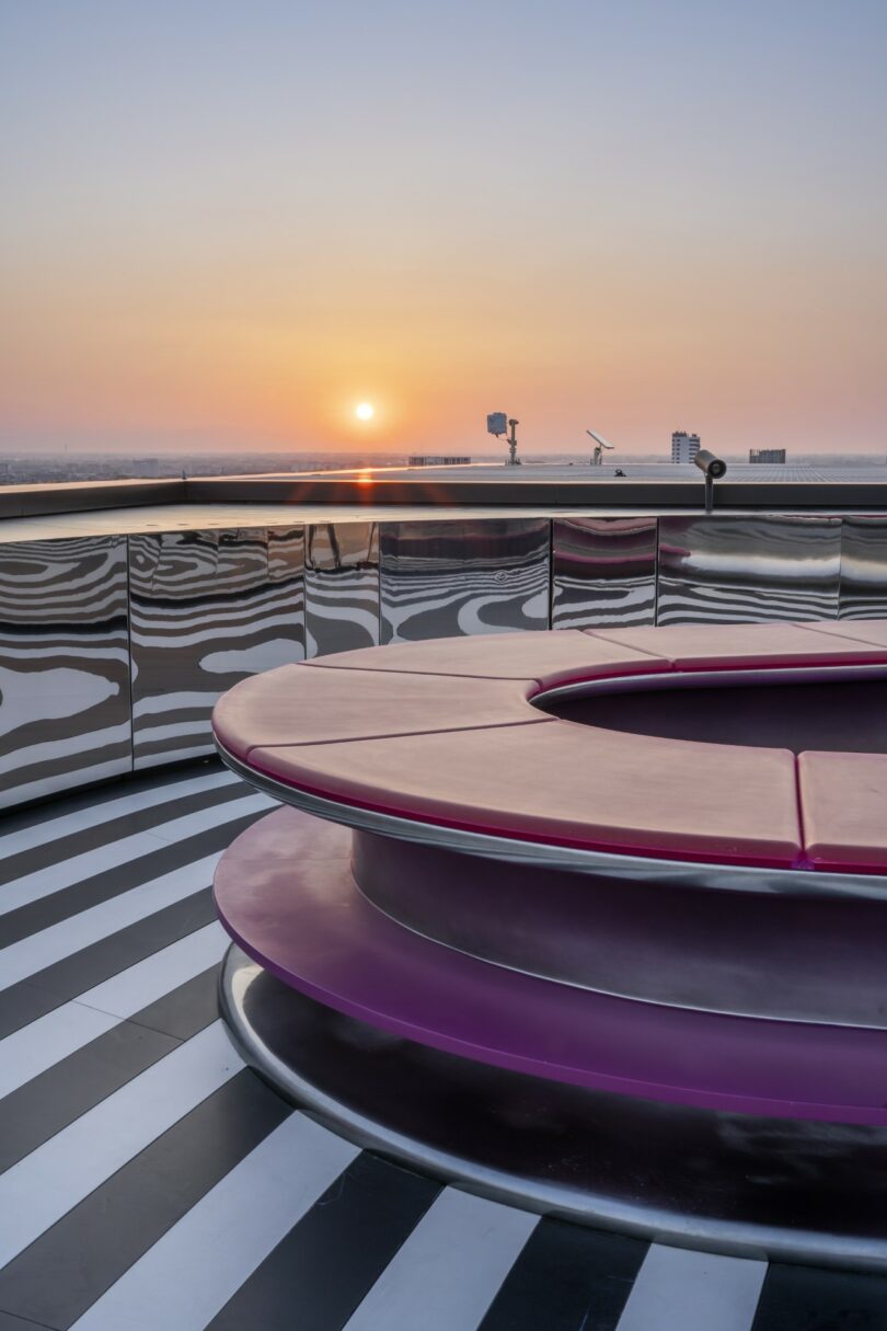 A round magenta bench on a black and white striped floor, with a cityscape and sunset visible in the background