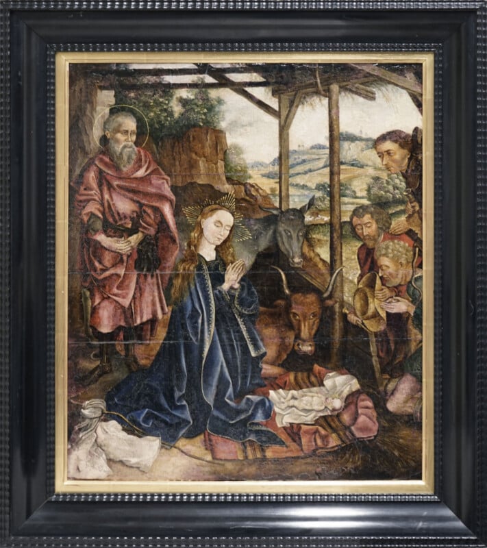 A framed painting of the Nativity shows Mary kneeling in prayer before baby Jesus in a manger, surrounded by Joseph, three wise men, an ox, and a donkey under a rustic shelter with fields in the background.