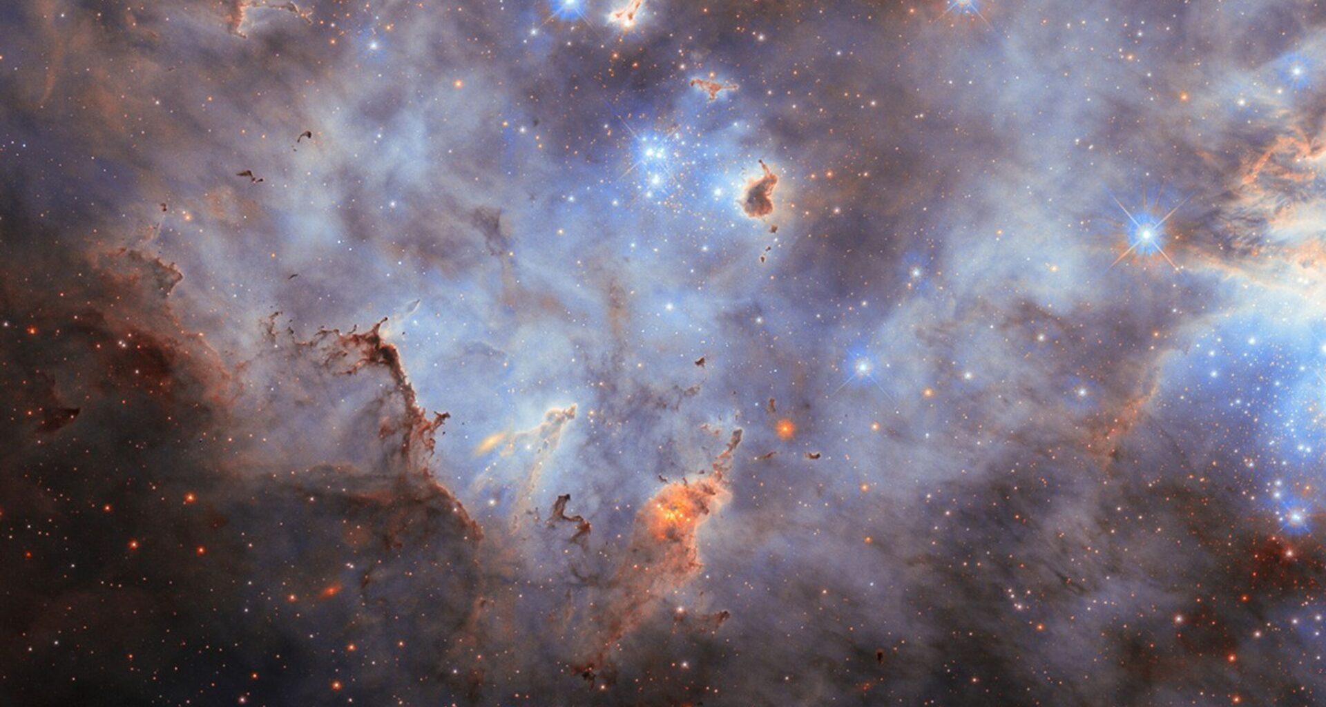 Stars in a star cluster shine brightly blue, with four-pointed diffraction spikes radiating from them. The center shows a small, crowded group of stars while a larger group is partially visible on the right side of the image. The nebula is mostly thick, smoky clouds of gas, lit up in blue tones by the stars. Clumps of dust hover before and around the stars; they are mostly dark but lit around their edges where the starlight erodes them.