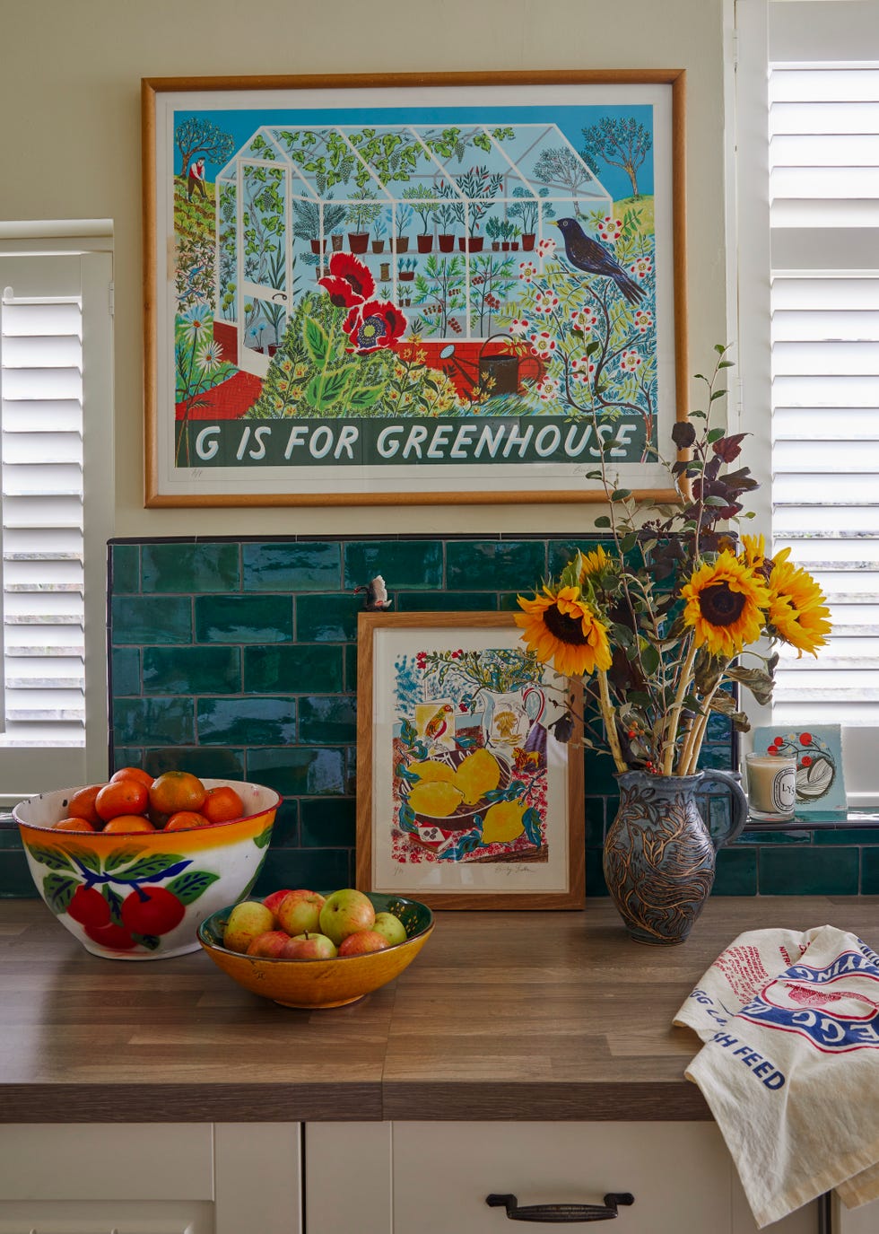 Emily Sutton’s York kitchen with artwork and sunflowers kitchen vignette in emily sutton’s york home with colourful framed prints, green tiled splashback, fruit bowls, and a jug of sunflowers