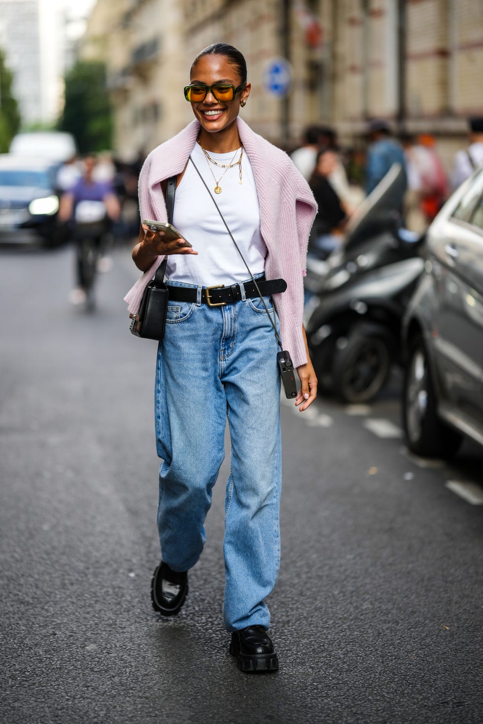 paris, france june 21: a guest wears orange sunglasses, gold earrings, a white halter neck tank top, gold chain pendant necklaces, a pale pink wool pullover, a black shiny leather shoulder bag from givenchy, a black crossbody camera, a black shiny leather belt, blue faded denim large mom jeans pants, black shiny leather loafers, during the bluemarble show, during paris fashion week menswear spring summer 2023, on june 21, 2022 in paris, france. (photo by edward berthelot/getty images )