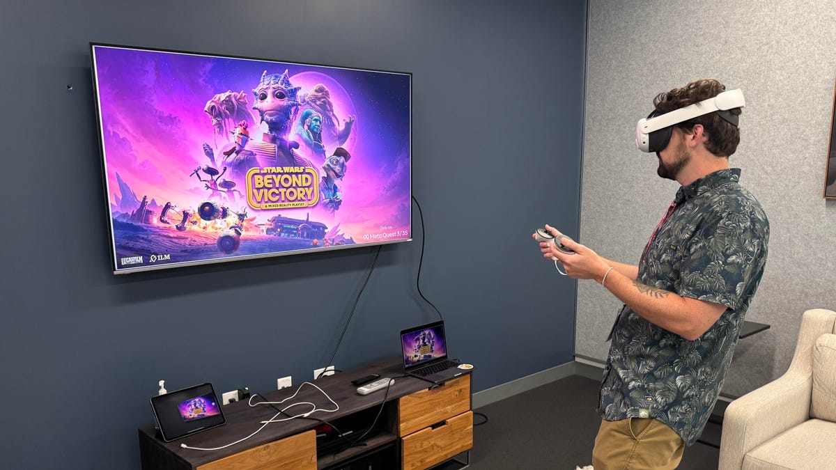 A man wearing a VR headset and holding controllers stands in front of a TV screen with the game&apos;s logo.