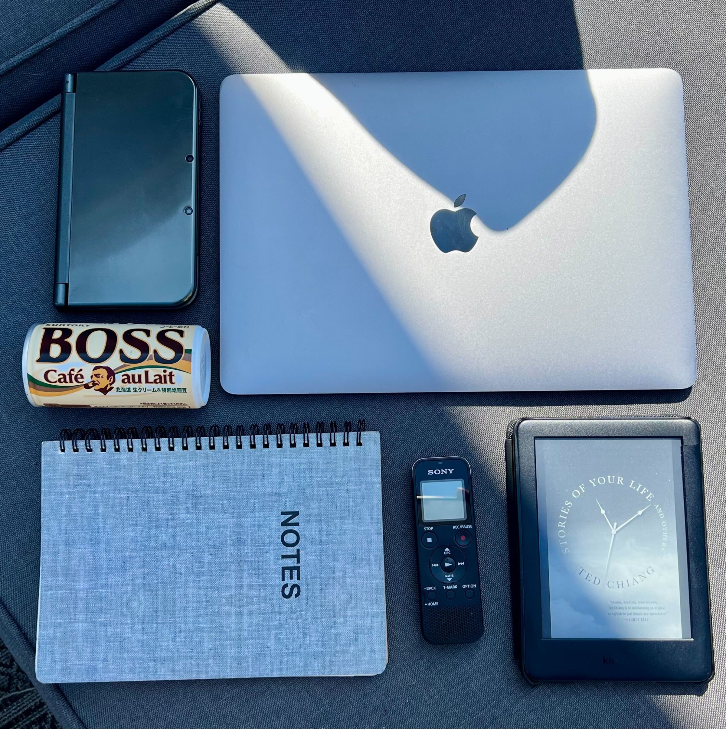 A photo of a laptop, Nintendo 3DS, Kindle, notebook, voice recorder, and can of Boss coffee.