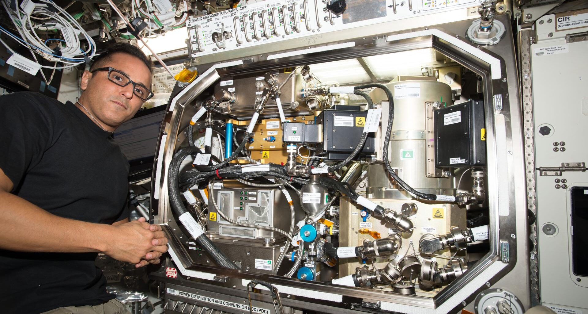 Astronaut in glasses working with scientific equipment inside an International Space Station laboratory.
