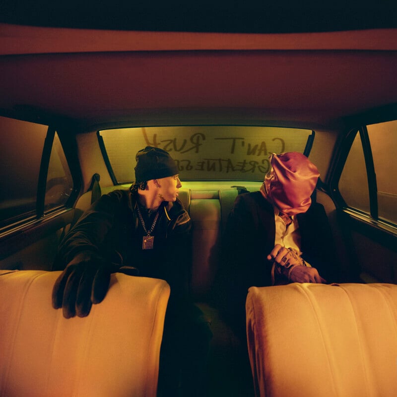 Two people sit in the back seat of a car at night. One wears dark clothes and a beanie, looking at the other, who is dressed in a suit with a pink mask covering their face. "CAN'T RUSH GREATNESS" is written on the foggy rear window.