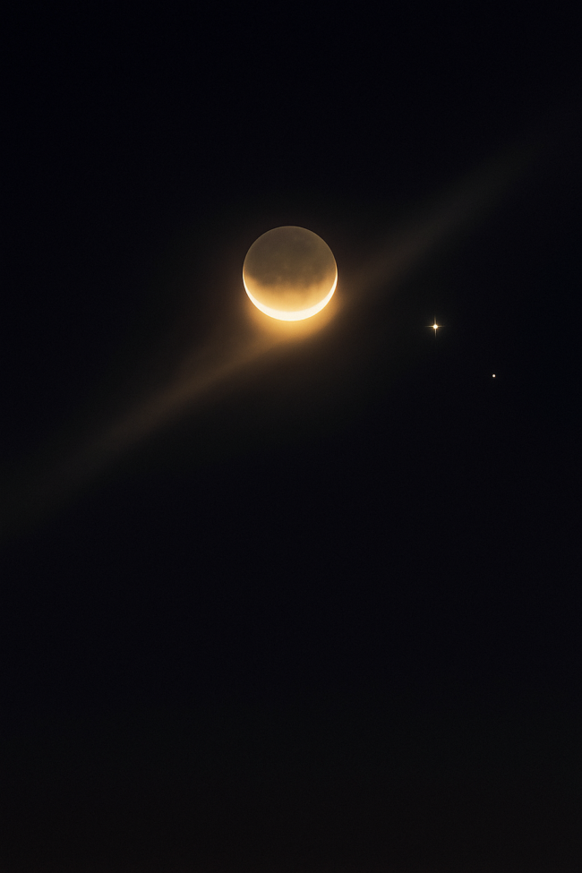 A yellowish glow on the bottom edge of a crescent moon with a bright and dim light nearby.