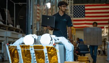 A person wearing glasses and a dark polo shirt stands indoors holding an open laptop in one hand. In front of him is a piece of large mechanical equipment resembling a planetary surface rover, with white structural arms and panels covered in gold foil. Behind him, another person is carrying a large, flat, rectangular object. The background shows industrial equipment and a large American flag hanging on the wall.