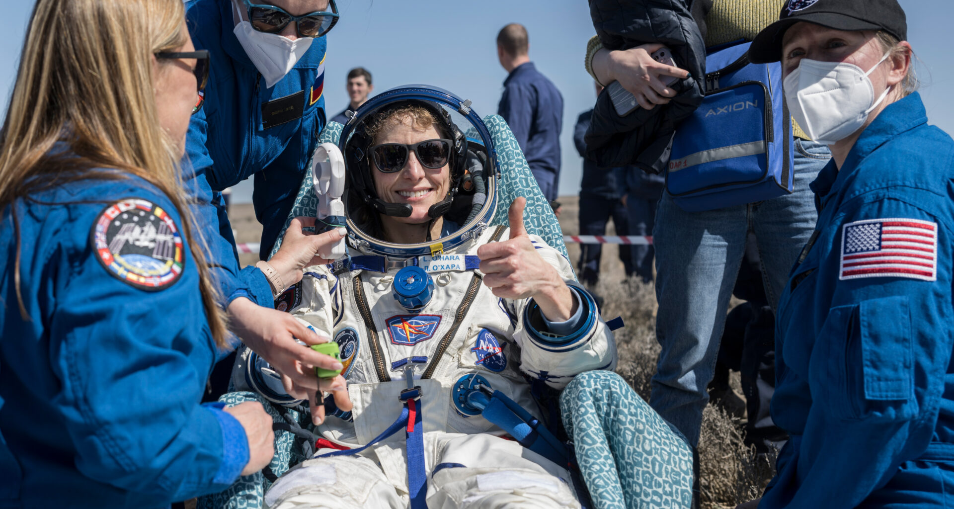 Three people in blue flight suits stand around a woman in a white space suit who