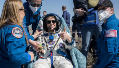 Three people in blue flight suits stand around a woman in a white space suit who