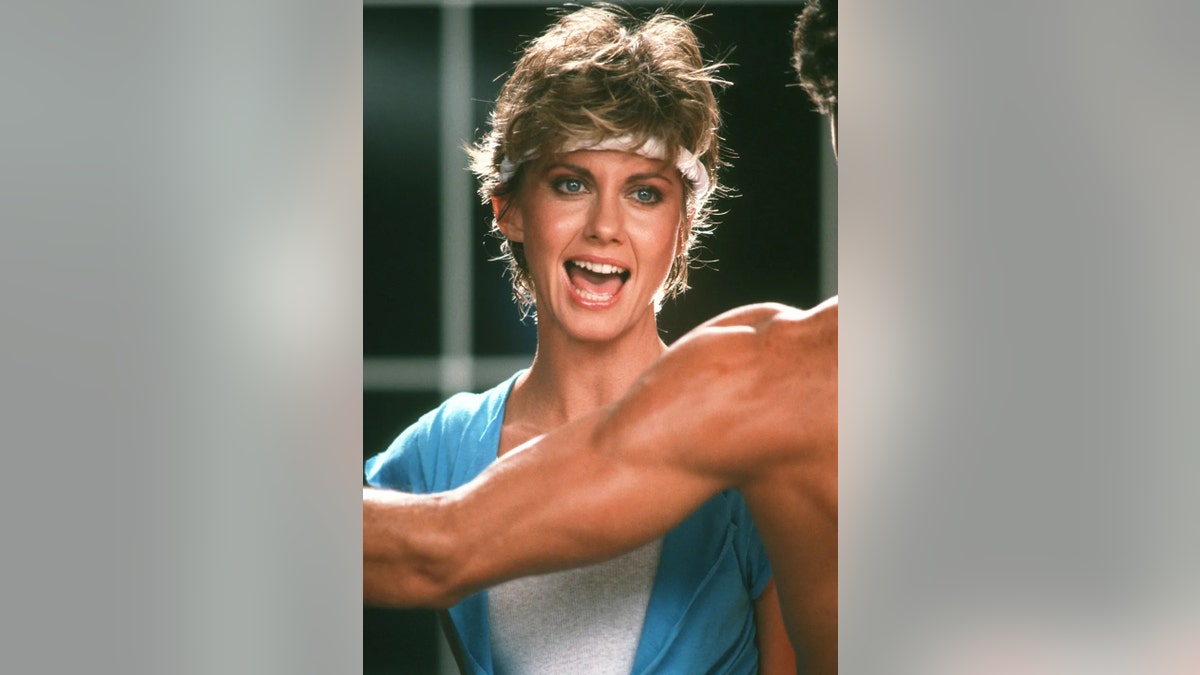 Olivia Newton-John singing in front of a muscled arm.