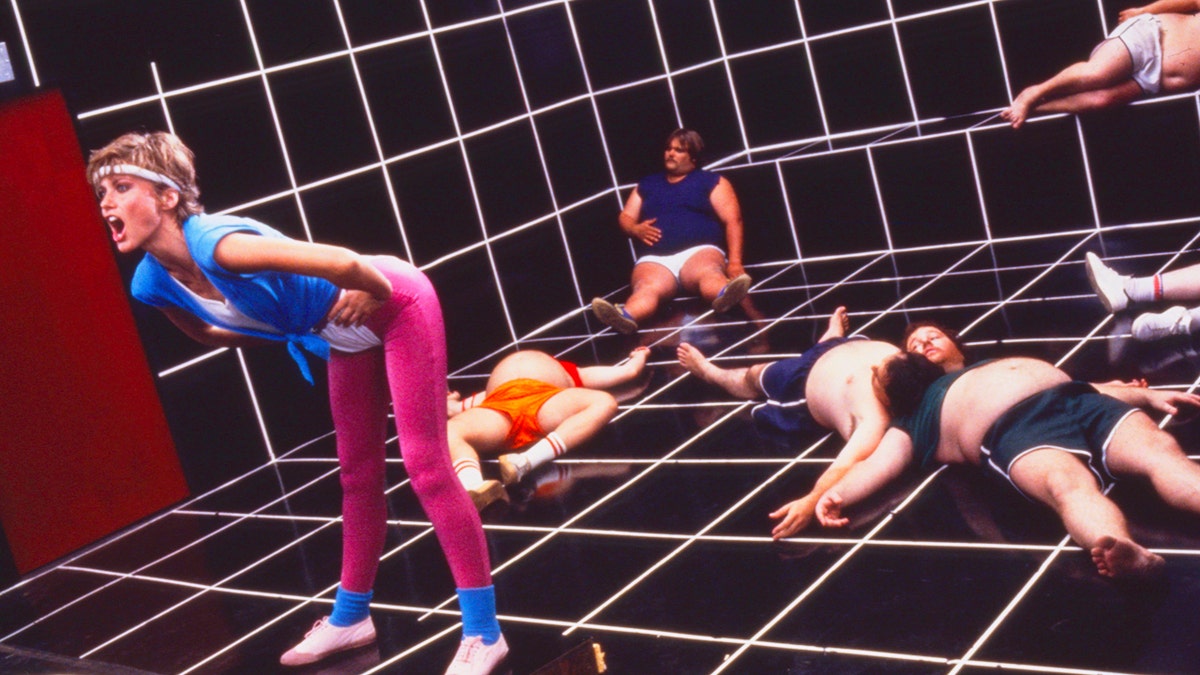 Olivia Newton-John performing "Physical" in front of a group of men in workout gear looking exhausted.