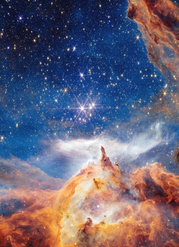 A colorful nebula in deep space with bright stars shining amidst swirling clouds of orange, yellow, and blue gas and dust, set against a dark blue star-filled background.