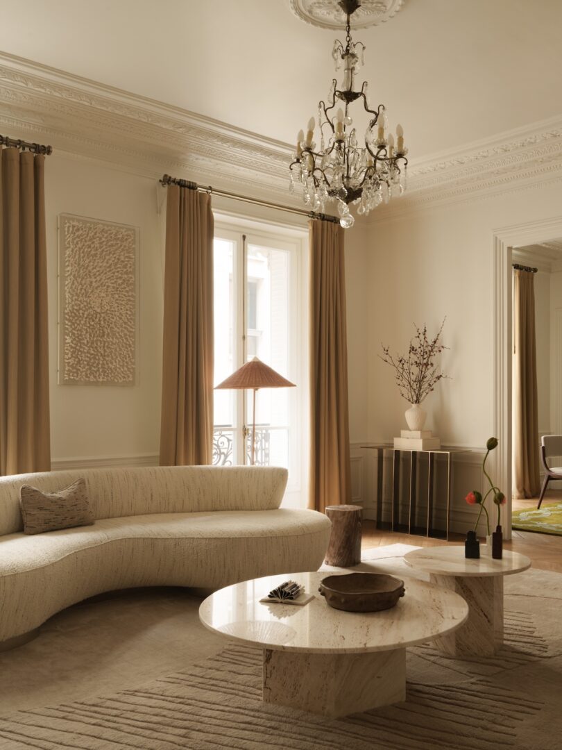 Elegant living room with a curved cream sofa, two round marble tables, beige curtains, a chandelier, and neutral-toned decor, including a lamp and a vase with branches.