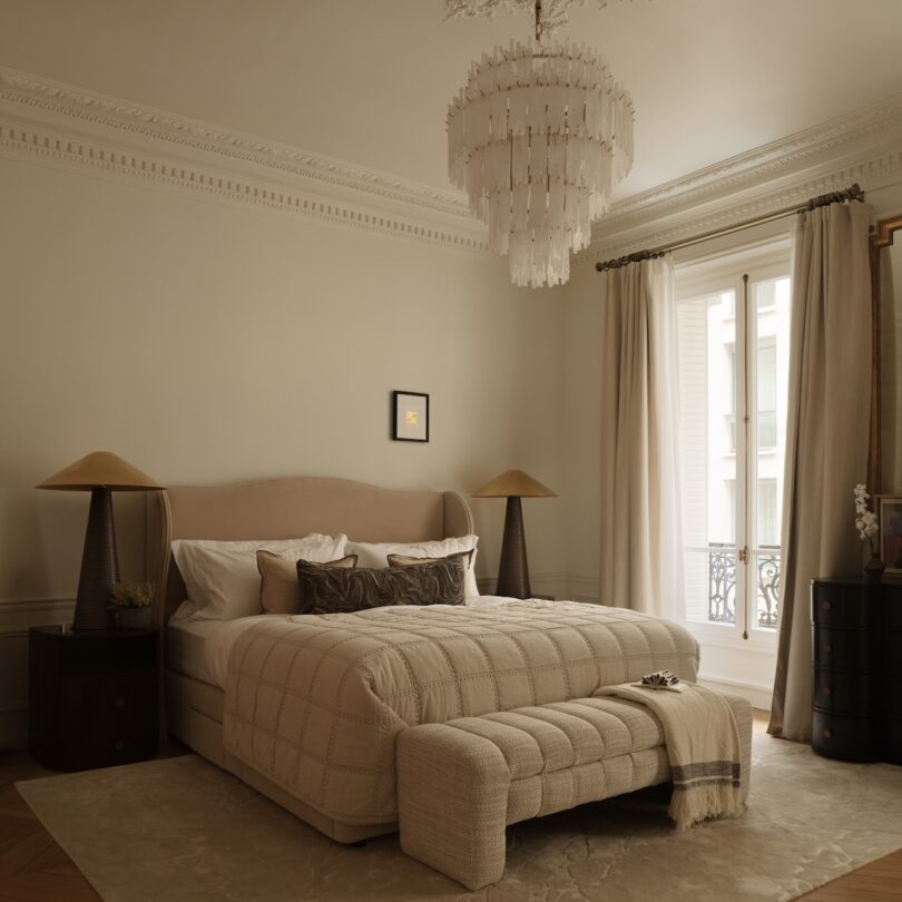 A bedroom with a beige upholstered bed, matching bench, two brown nightstands with lamps, a chandelier, large window with curtains, and light-colored walls and rug.