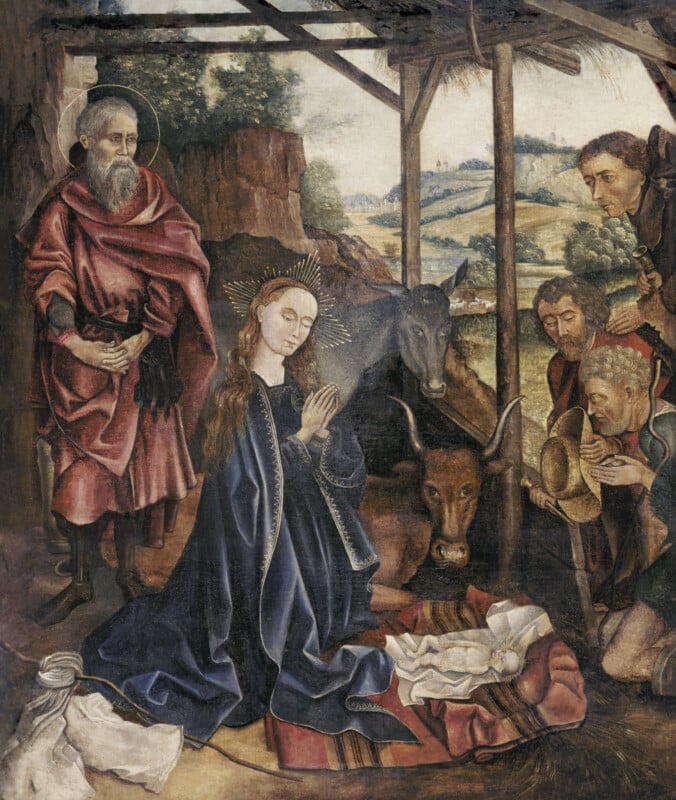 A nativity scene with Mary praying beside baby Jesus in a manger, surrounded by Joseph, shepherds, an ox, and a donkey, set in a rustic stable with a landscape visible in the background.
