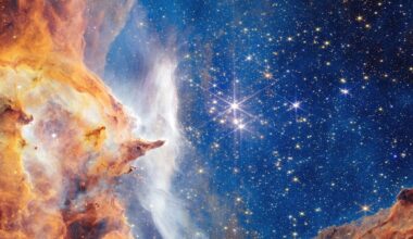 A vibrant image of outer space showing a bright star cluster surrounded by clouds of orange, yellow, and white gas and dust, with a dark blue background filled with many sparkling stars.