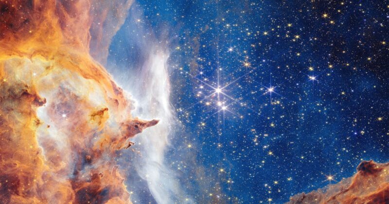 A vibrant image of outer space showing a bright star cluster surrounded by clouds of orange, yellow, and white gas and dust, with a dark blue background filled with many sparkling stars.