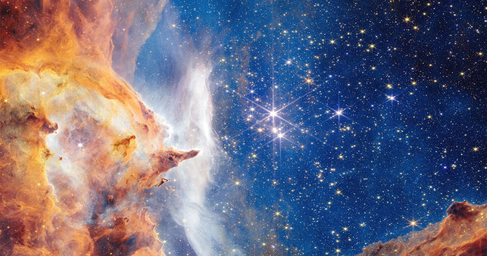 A vibrant image of outer space showing a bright star cluster surrounded by clouds of orange, yellow, and white gas and dust, with a dark blue background filled with many sparkling stars.