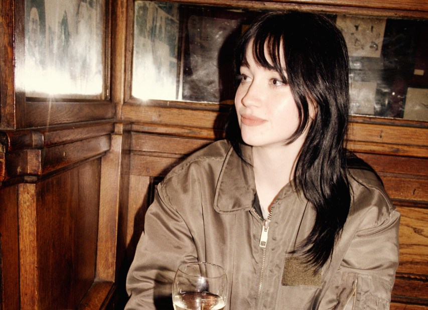 A woman in a dark green jacket sits at a wooden table with a glass of white wine and two glasses of beer.
