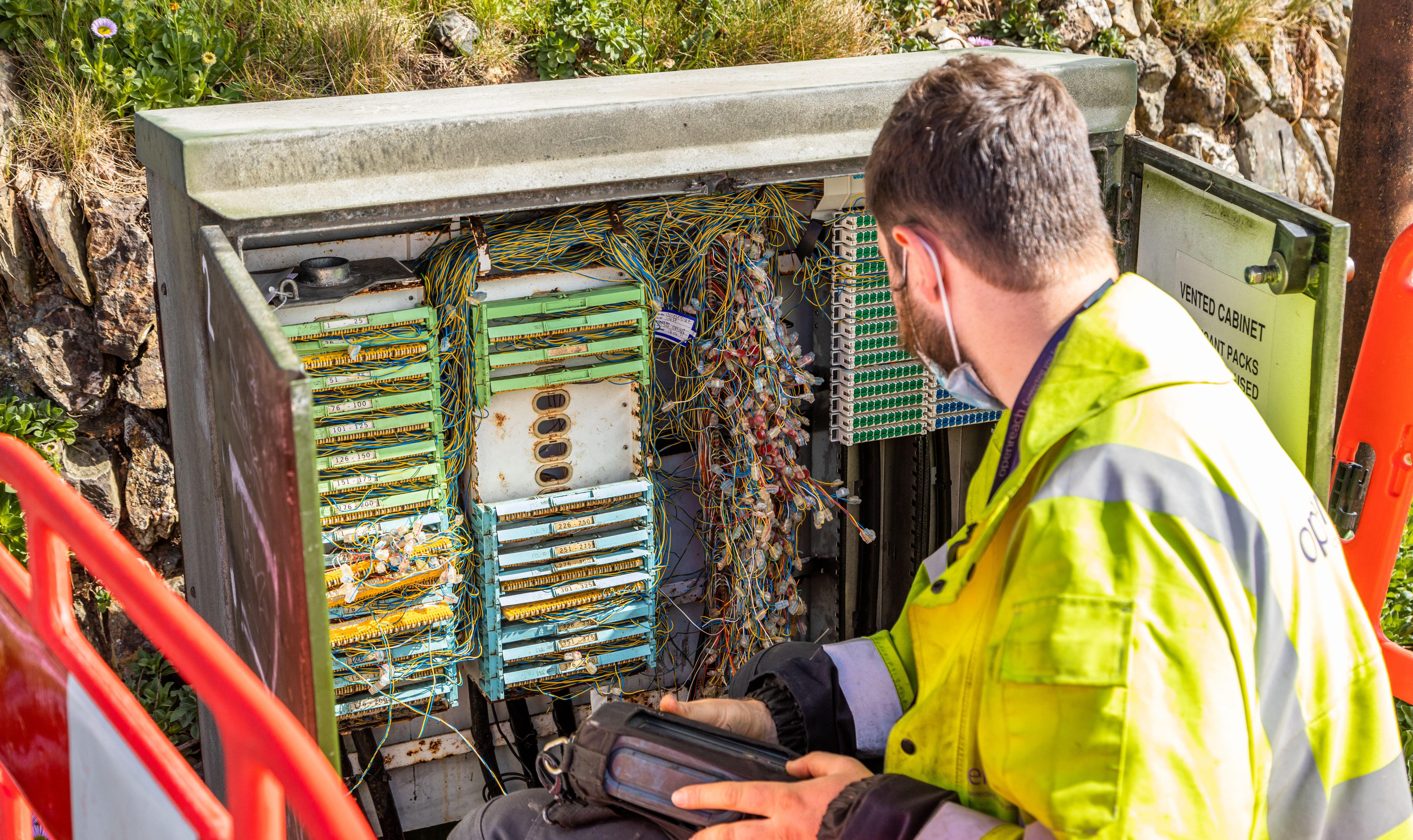 A telecoms engineer in a high-visibility jacket and face mask works on solving connection issues in an open street cabinet filled with numerous wires.