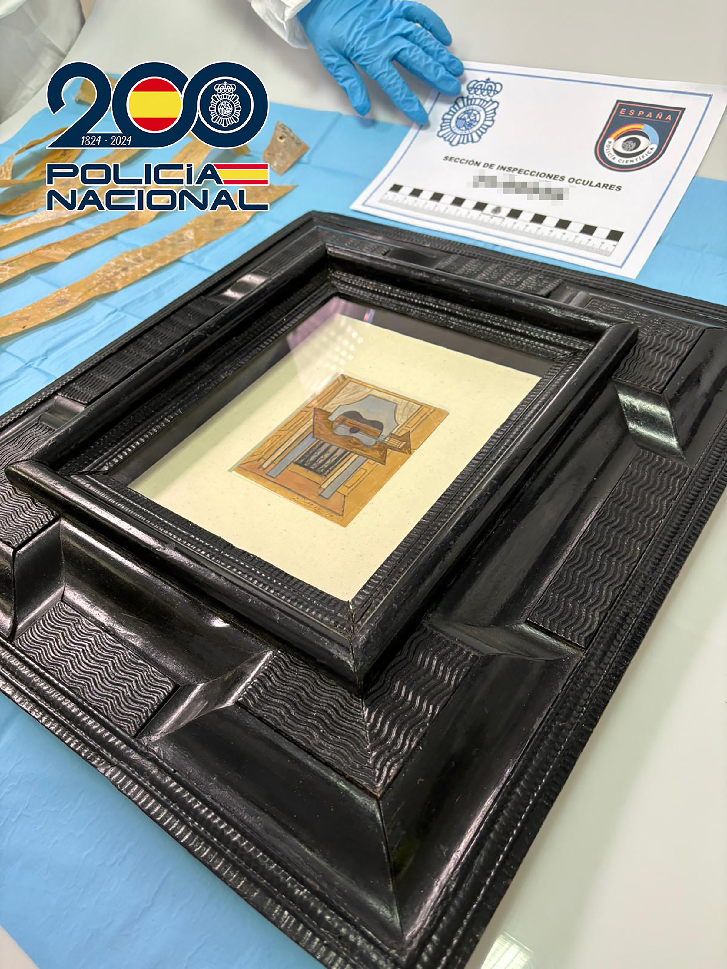 Gouache and pencil painting "Still Life with Guitar" by Pablo Picasso, framed in black, on a blue cloth alongside a Spanish National Police document.