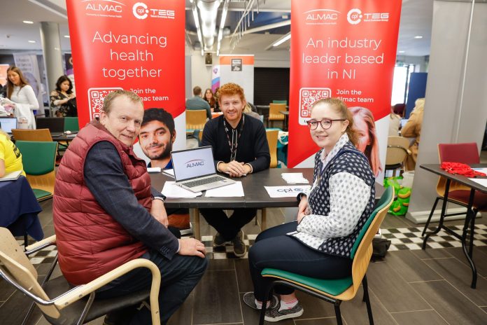 Three people sitting at a job fair stand for Almac in Portadown, surrounded by branded signs.