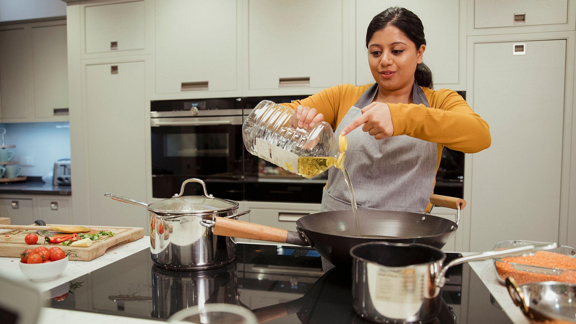 A stock photo shows a woman pouring a big bottle of sunflower oil into a deep wok pan on an electric hob with cooking ingredients all around her and her kitchen cabinets and oven in the background.