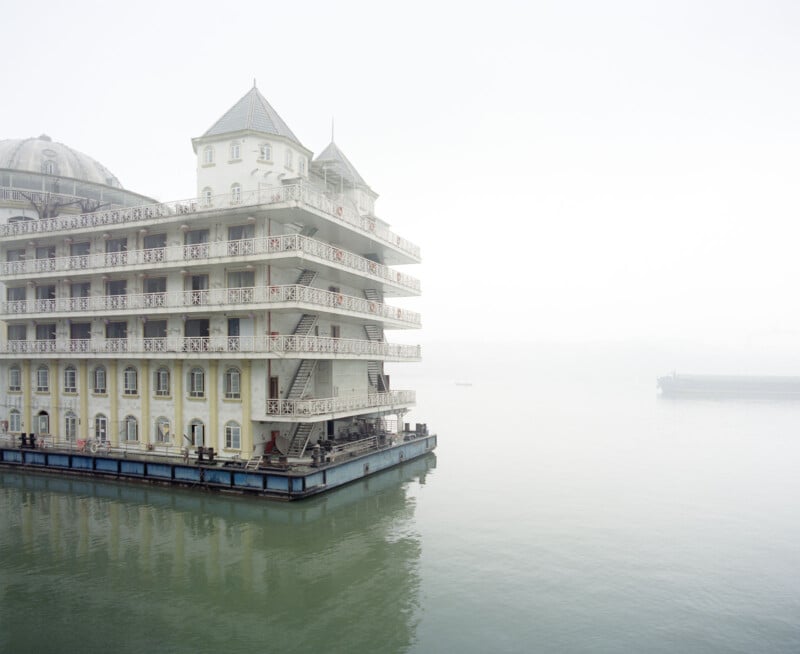 A large, ornate, multi-story floating building resembling a historic hotel sits on calm, foggy water with faint structures visible in the misty background.
