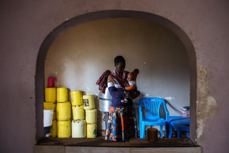 A woman stands holding a baby inside a room, viewed through an arched opening. Yellow water containers and blue plastic chairs are against the wall behind her. The setting appears modest and simple.