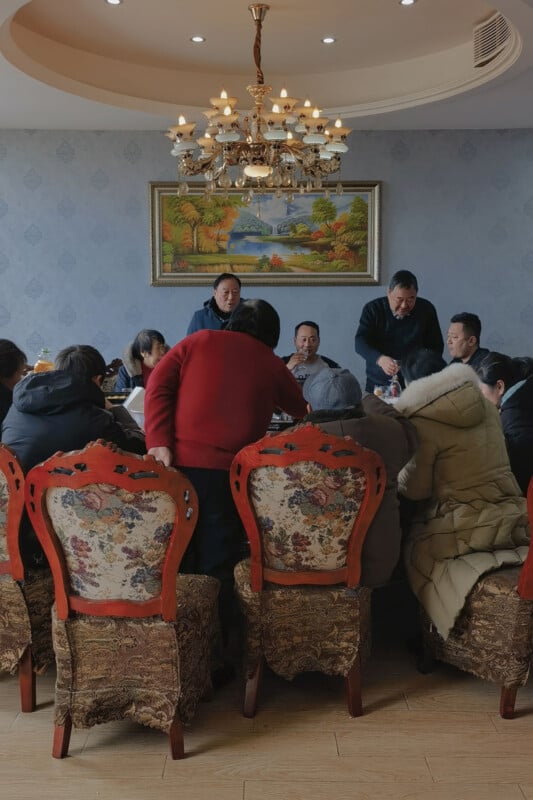 A group of people sit around a dining table in a warmly lit room with ornate chairs, sharing a meal. A chandelier hangs above, and a landscape painting decorates the wall behind them.