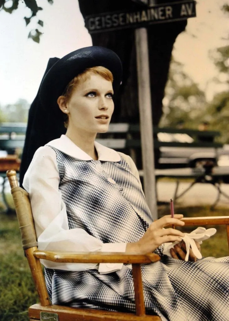 Person seated outdoors, wearing a checkered dress with a wide-brimmed hat, holding gloves and a pencil near a street sign