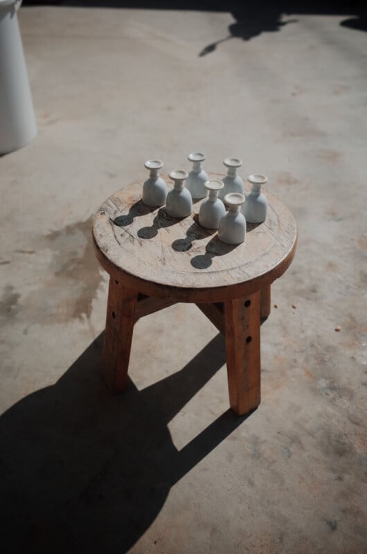 A round wooden stool with seven small white ceramic vases arranged on top, casting shadows on the stool and concrete floor in bright sunlight.