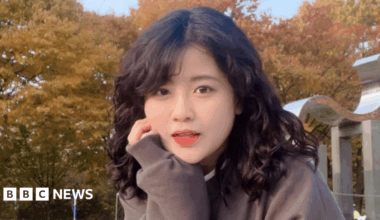 Baek Se-hee resting her cheek on her hand as she looks at the camera. She has curly black hair and is wearing a brown sweater. In the background are trees with yellow leaves.