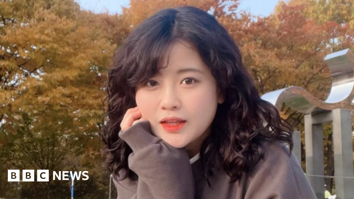 Baek Se-hee resting her cheek on her hand as she looks at the camera. She has curly black hair and is wearing a brown sweater. In the background are trees with yellow leaves.