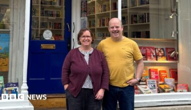 Gideon and Cath York standing beside each other outside their shop,  Cotswold Book Room. Cath is wearing black jeans, a black and white spotted blouse beneath a dark purple cardigan, and has short brown curly hair and glasses. Gideon is bald and is wearing a yellow t-shirt with dark blue jeans. The shop has a dark blue front door and in the windows there are lots of books on display, lit up with warm spotlights.