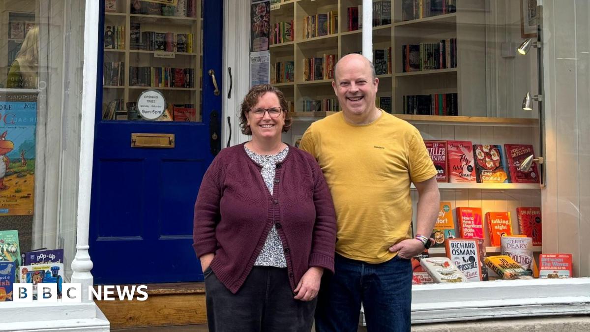 Gideon and Cath York standing beside each other outside their shop,  Cotswold Book Room. Cath is wearing black jeans, a black and white spotted blouse beneath a dark purple cardigan, and has short brown curly hair and glasses. Gideon is bald and is wearing a yellow t-shirt with dark blue jeans. The shop has a dark blue front door and in the windows there are lots of books on display, lit up with warm spotlights.