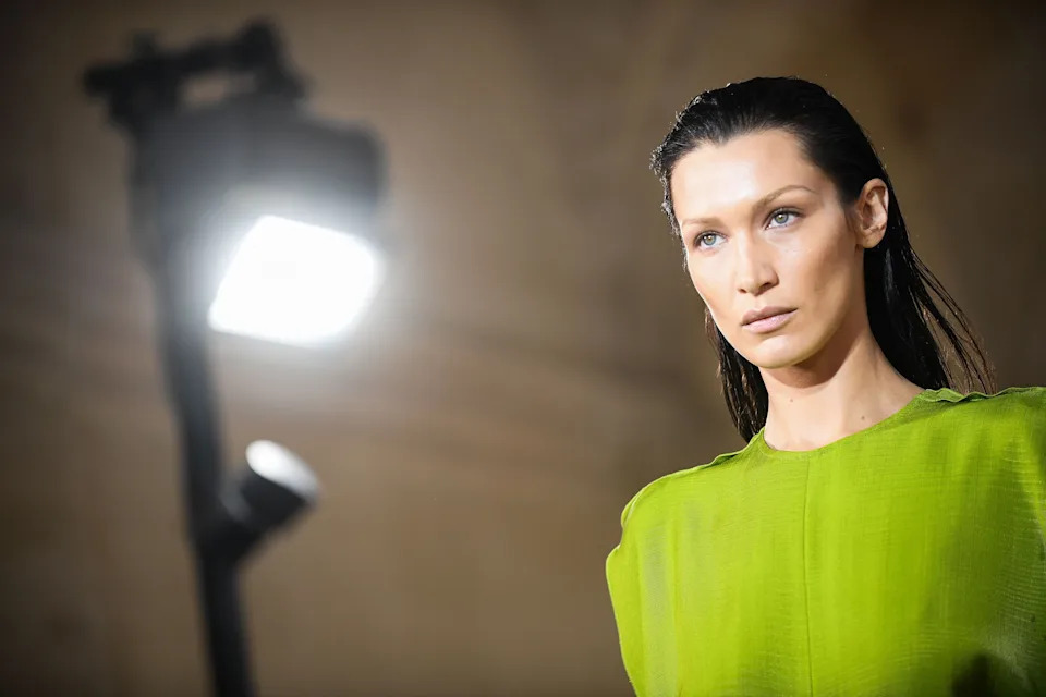 Bella Hadid with wet slicked-back hair wearing a structured, long-sleeve garment on the runway, with bright overhead lighting