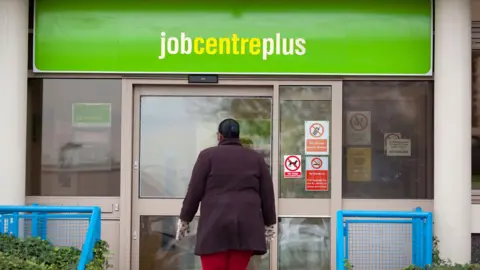 Alamy A stock image shows a woman walking into a Job Centre plus. She is wearing a purple knee length coat with red throusers. Above her, the words job centre plus are written in white and yellow against a green background.