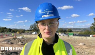 Jack Little on a construction site, wearing a high vis jacket, a blue hard hat, and protective glasses.