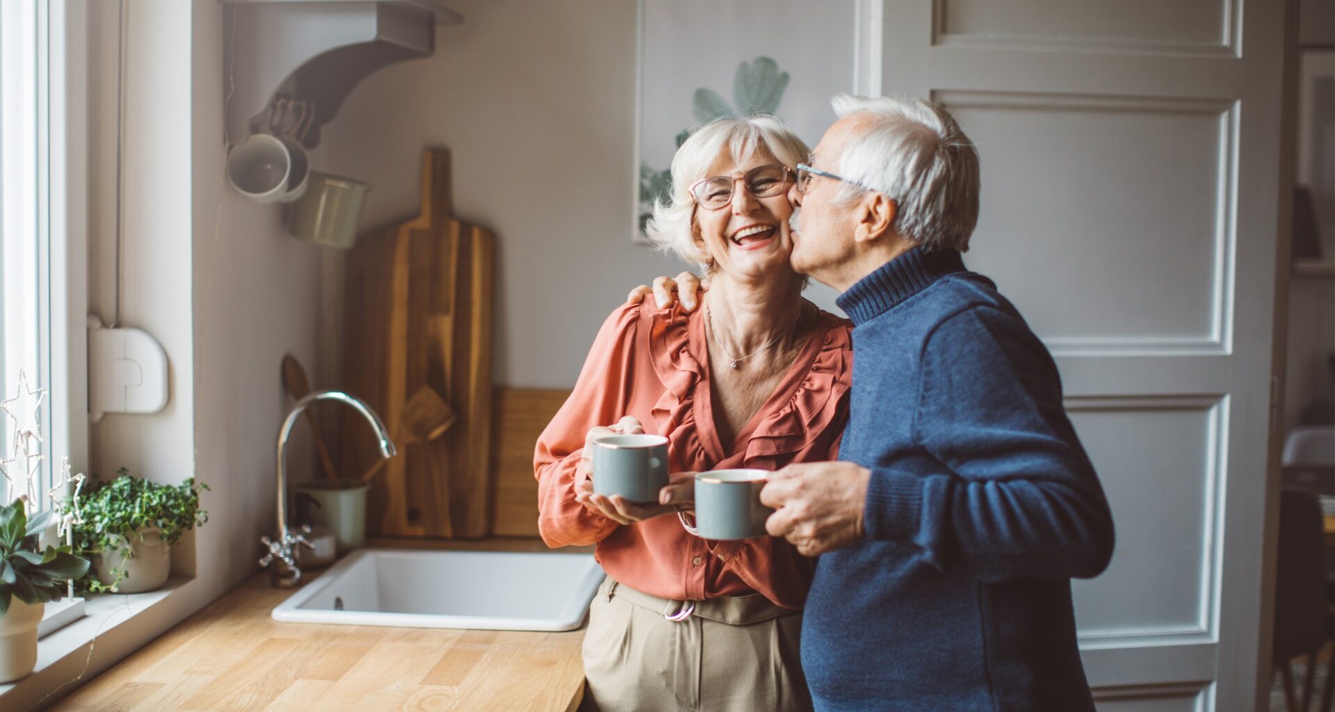 A happy retired couple stands in their kitchen as the husband give his wife a kiss on the cheek.