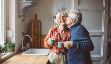 A happy retired couple stands in their kitchen as the husband give his wife a kiss on the cheek.